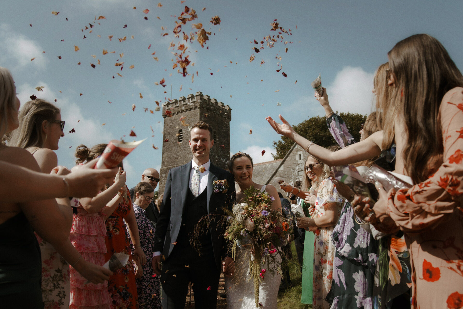 Wedding guests throwing flower petals at smiling bride and groom on a sunny day.