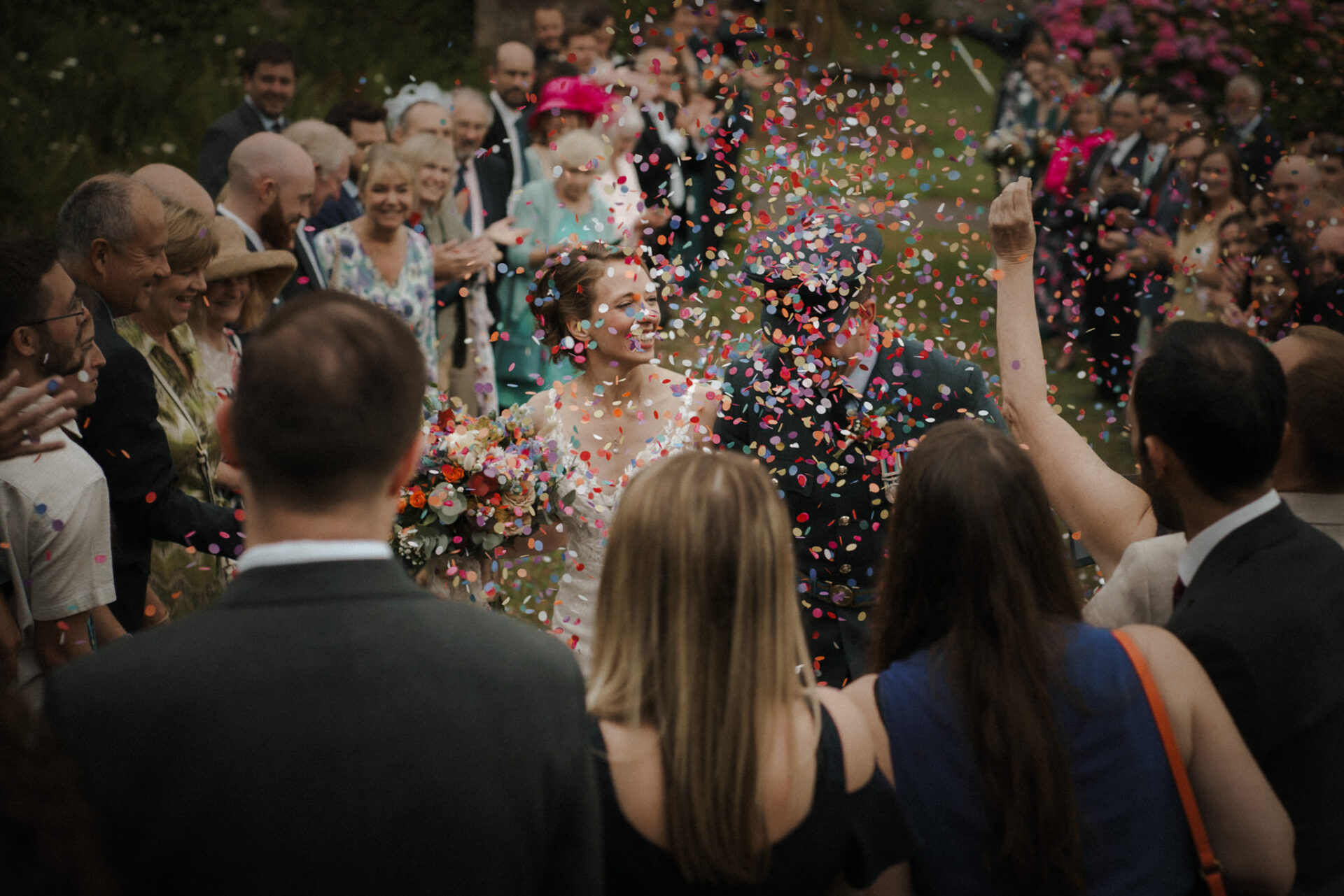 A bride and groom walk through a crowd as guests toss colorful confetti at a wedding celebration.