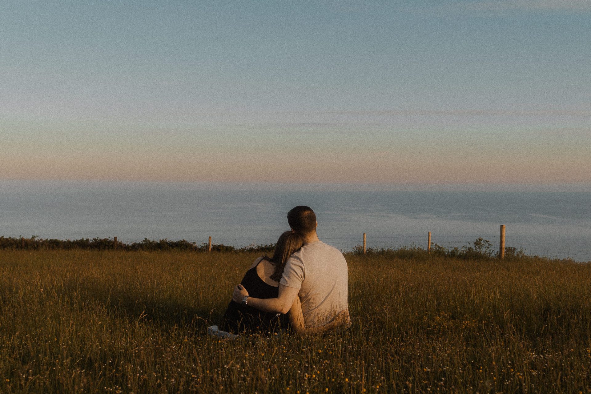 A couple sitting on a grassy hill overlooking the ocean.