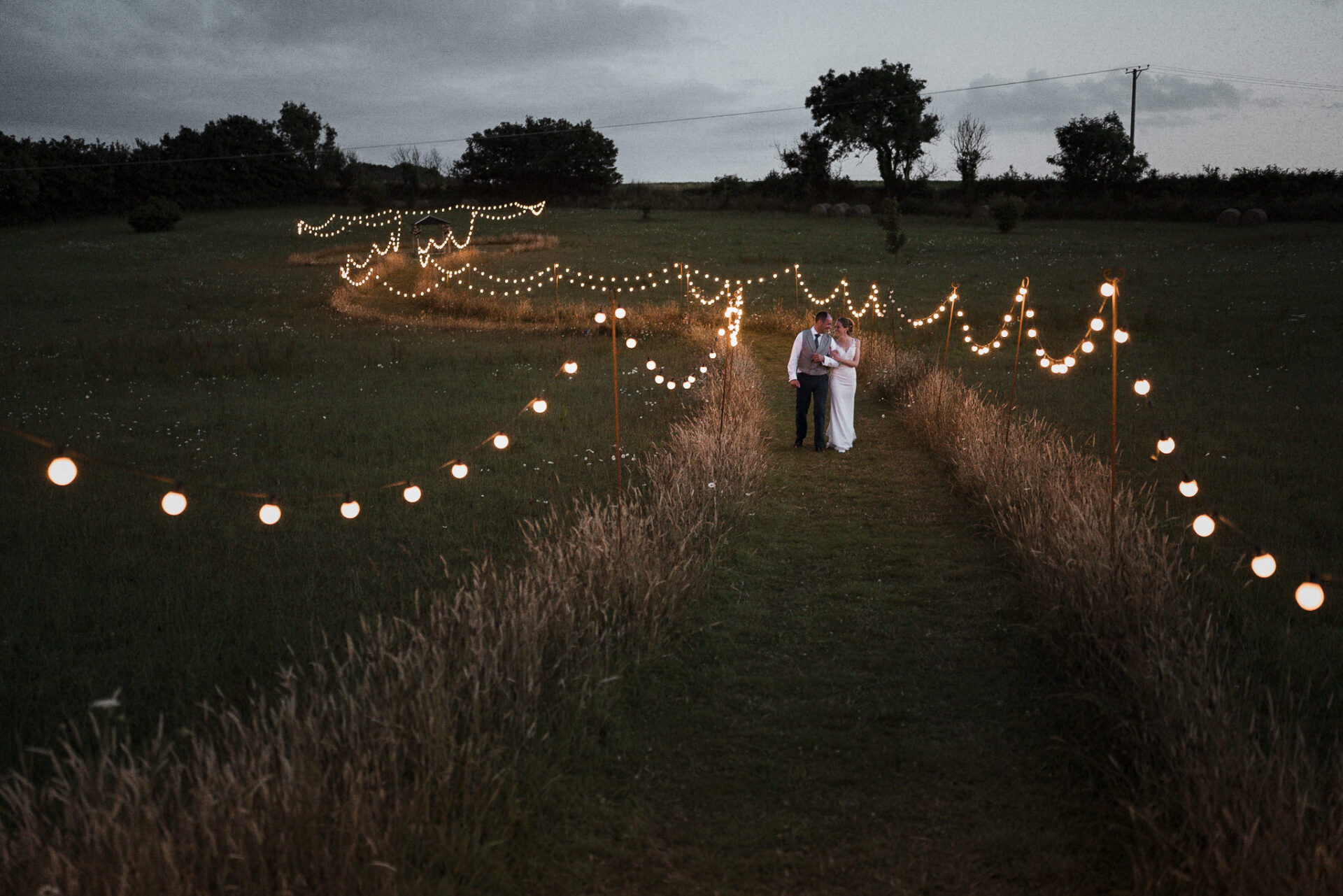 A recent wedding with a bride and groom standing in a field with string lights.
