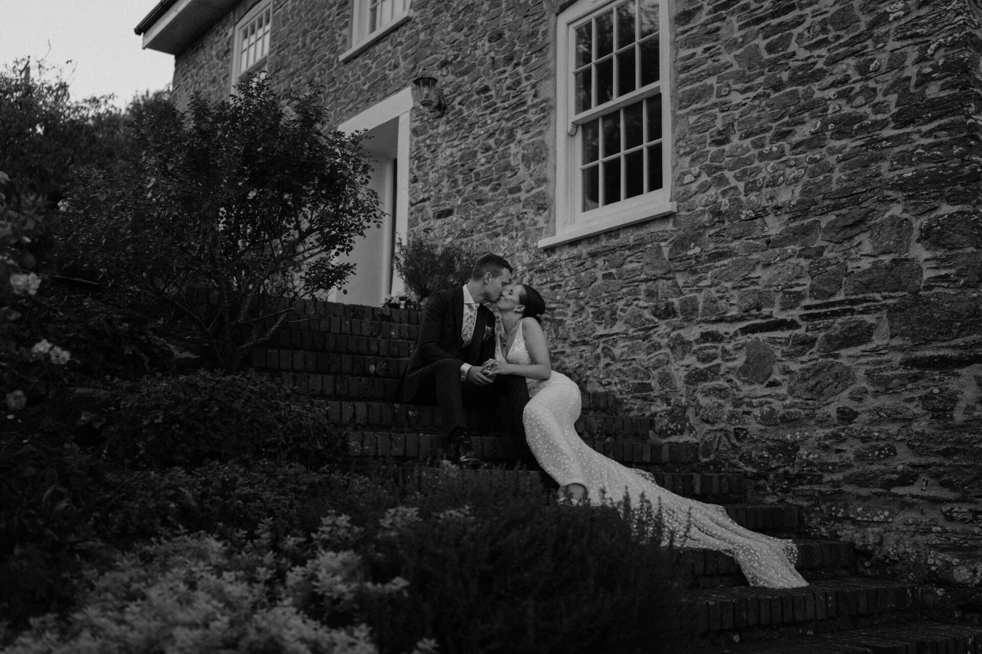 A bride and groom sitting on steps in front of a stone house.